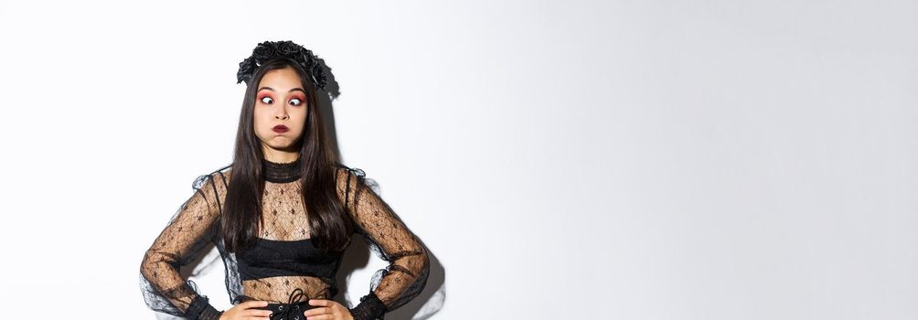 Portrait of funny and carefree asian girl having fun on halloween, making faces and holding breath, standing in gothic witch costume over white background.