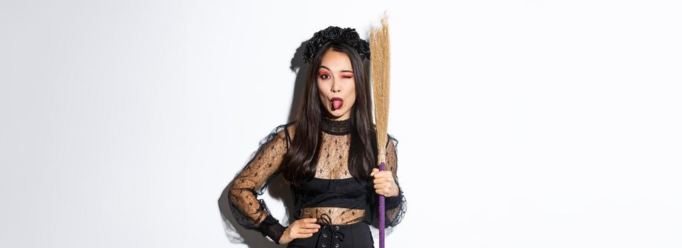 Beautiful asian woman celebrating halloween in witch costume, holding broom and showing tongue, winking silly at camera, standing over white background.