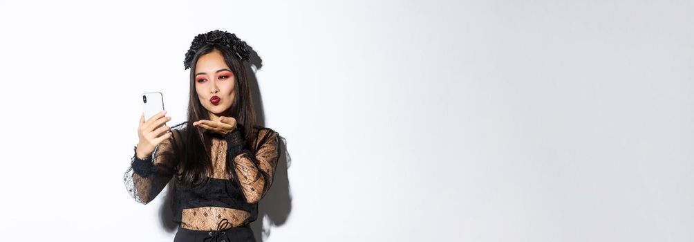 Portrait of stylish asian female blogger with gothic makeup and halloween costume sending air kiss at mobile phone camera, record video or having videocall, standing over white background.