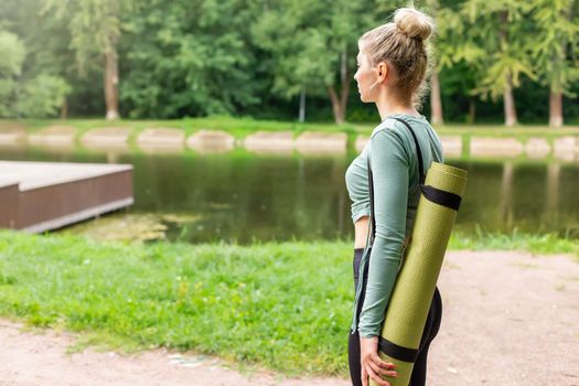 A slender blonde with a green sports mat behind her back is walking in a summer park. Side view. To close. Close up. Copy space