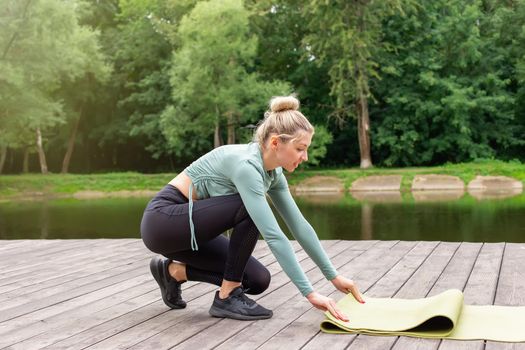 A woman in sportswear, in a green top and black leggings, in the summer by pond, unwinds a green gym mat, prepares for sports. side view. Copy space