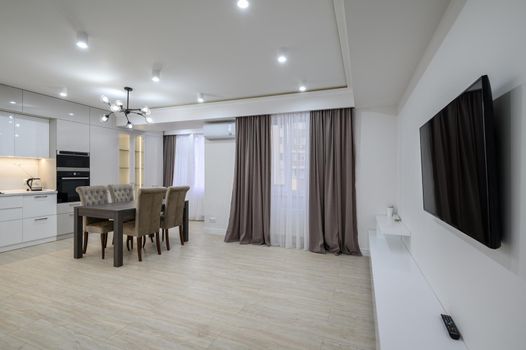 Interior of a huge white luxury kitchen in a studio apartment, tv set, lots of copy space, large windows with curtains and a dining table with chairs