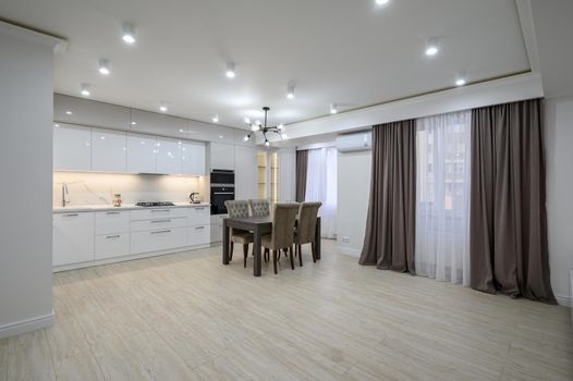 Interior of a huge white luxury kitchen in a studio apartment, lots of copy space, large windows with curtains and a dining table with chairs