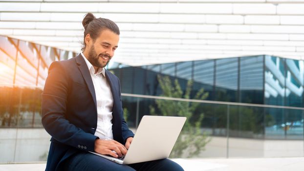 Business on the Go Businessman Working on his Laptop on the Street near office building. Handsome hispanic business person sitting use laptop. Confident male manager working outdoor. Banner size.