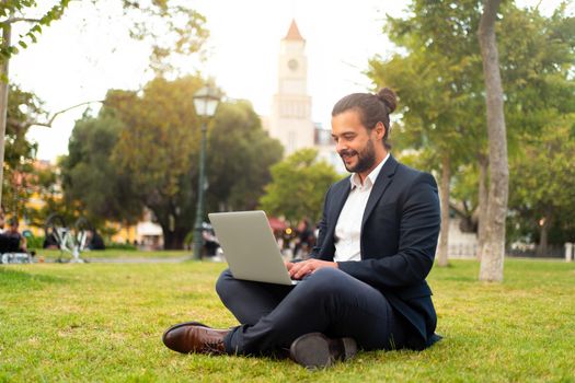Handsome hispanic male businessman sitting lotus position in public park use laptop. Remote work concept. Stylish european man in suit relax in park summer day working on computer. Technology.