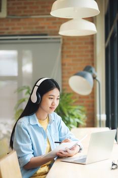 Happy young asian woman sitting at table and studying and learning using laptop. Student learning and studying online using laptop sitting at table and wearing headset. Online learning concept.