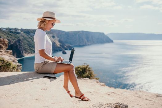 Successful business woman in yellow hat working on laptop by the sea. Pretty lady typing on computer at summer day outdoors. Freelance, travel and holidays concept.