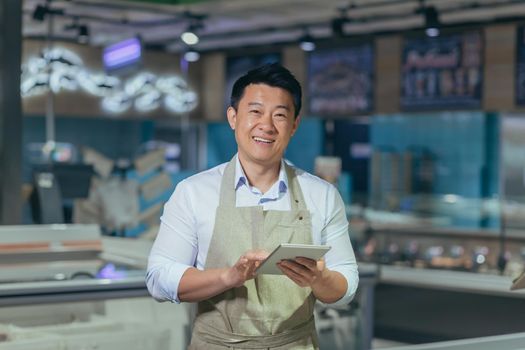 Portrait of a handsome young Asian man, an employee of a store, supermarket. Standing in an apron, holding a notepad, looking at the camera, smiling