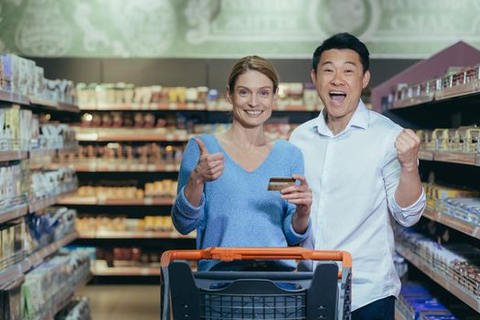 Family shopping. Young beautiful interracial couple shopping in a supermarket. Happy Asian woman and man standing with cart, showing credit card, satisfied with shopping, prices, discounts.