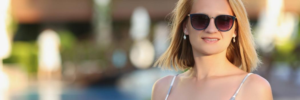 Portrait of lovely woman walking on hotel territory alone on summer evening. Blonde female in dress with resort on background. Holiday concept. Copy space