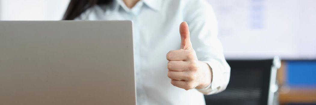 Close-up of office clerk showing thumbs up gesture working on laptop. Smiling employee express joy, good job, support business. Corporate, office concept