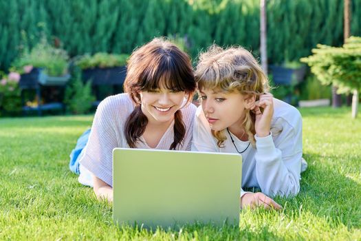 Two teenage girlfriends 17, 18 years old using laptop lying outdoors on grass on sunny summer day. Young female student looking at laptop screen, technology for leisure study entertainment