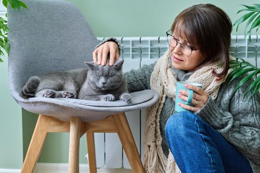 Cold winter season woman with cat near heating radiator. Middle-aged female in warm woolen knitted sweater with cup of hot drink and pet lying on chair. Heating season, lifestyle, crisis energy saving