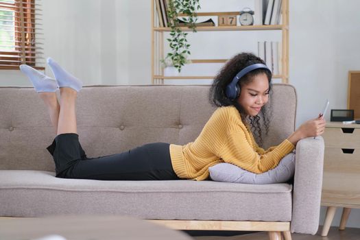african young woman relaxing at home lying on sofa and listening to music on tablet wearing headphones. girl relaxing on the sofa