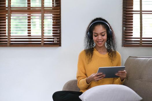 Photo of modern asian woman wearing headphones holding digital tablet while lying on sofa in home.