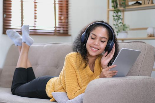 african young woman relaxing at home lying on sofa and listening to music on tablet wearing headphones. girl relaxing on the sofa of a cozy home.