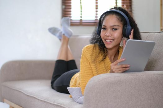 african young woman relaxing at home lying on sofa and listening to music on tablet wearing headphones. girl relaxing on the sofa of a cozy home.