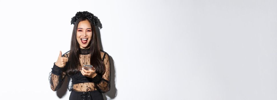 Portrait of satisfied asian woman in elegant gothic dress and black wreath showing thumbs-up while using mobile phone, standing over white background.