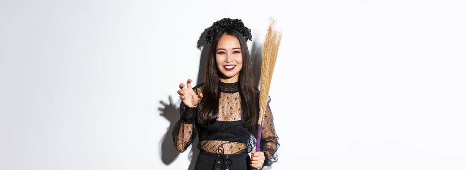 Image of beautiful asian woman in witch costume holding broom and smiling at camera, celebrating halloween, standing over white background.