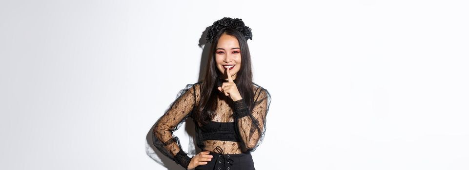 Smiling beautiful asian woman in black lace dress celebrating all saints day, have surprise for halloween, shushing with finger pressed to lips, share a secret, standing over white background.