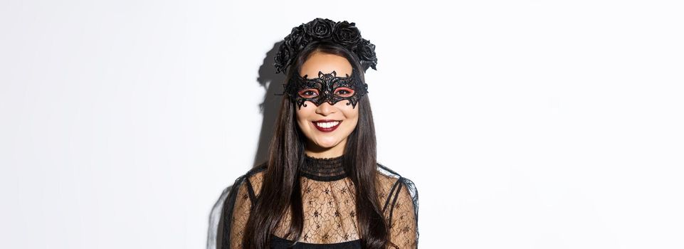 Close-up of mysterious woman in gothic wreath and black mask smiling at camera, celebrating halloween, standing over white background.