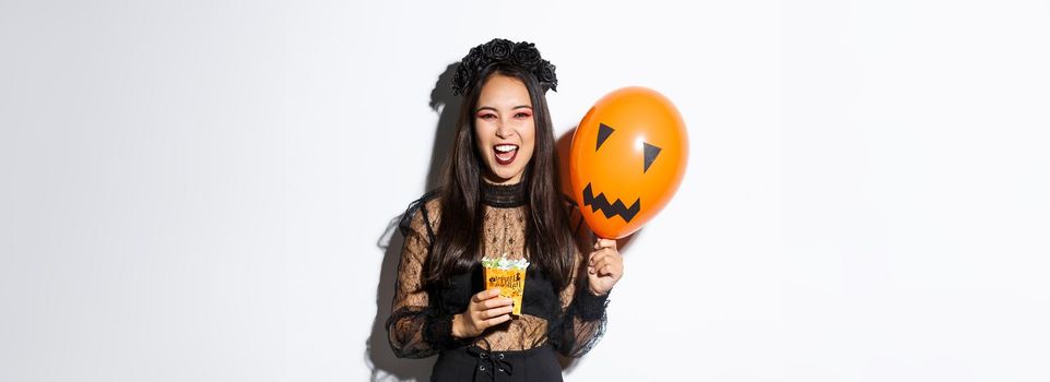 Scary witch trick or treating on halloween, holding sweets and orange balloon, standing in gothic lace dress with black wreath.