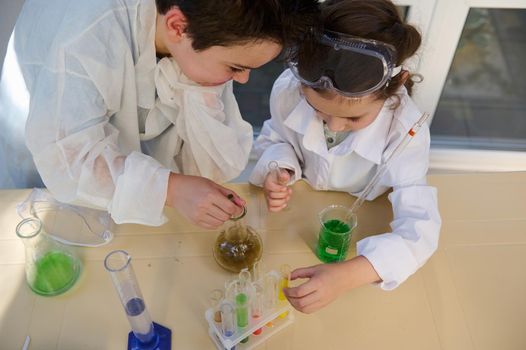 Overhead view of smart school kids, wearing white lab coat, learning Chemistry in the school science laboratory, conducting fascinating chemical experiment, using regents, chemicals and glass labware