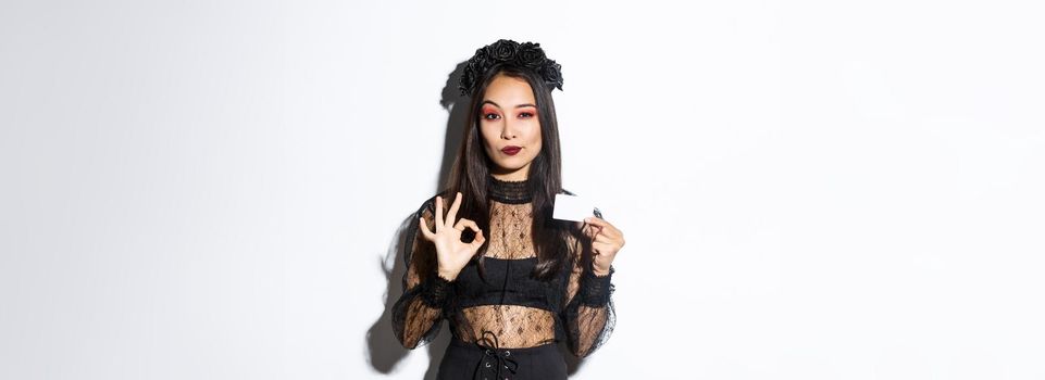 Portrait of confident asian woman assuring you in something, wearing halloween costume, showing okay gesture and credit card, white background.