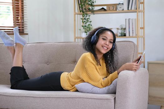 african young woman relaxing at home lying on sofa and listening to music on tablet wearing headphones. girl relaxing on the sofa of a cozy home.