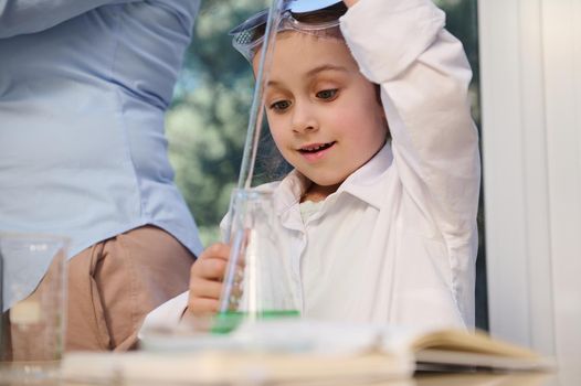 Smart Caucasian preschool little girl wearing white lab coat, learning Chemistry in the school science laboratory, conducting chemical experiment, using regents, chemicals and glass labware