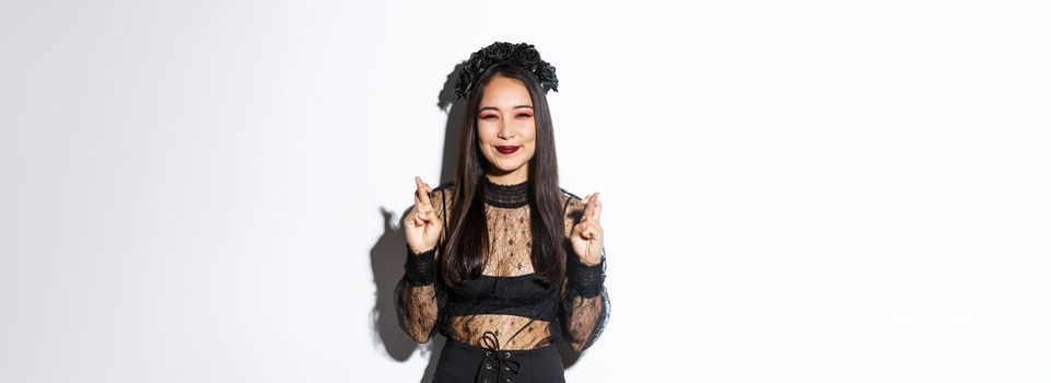 Image of happy asian girl who loves celebrating halloween dressed-up as witch, smiling hopeful and cross fingers for good luck, making wish, standing over white background.