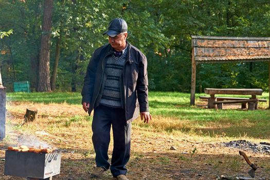 A engaging or ready to engage in physically energetic pursuits. An elderly cheerful man prepares a bonfire barbecue grill for cooking meat at a picnic