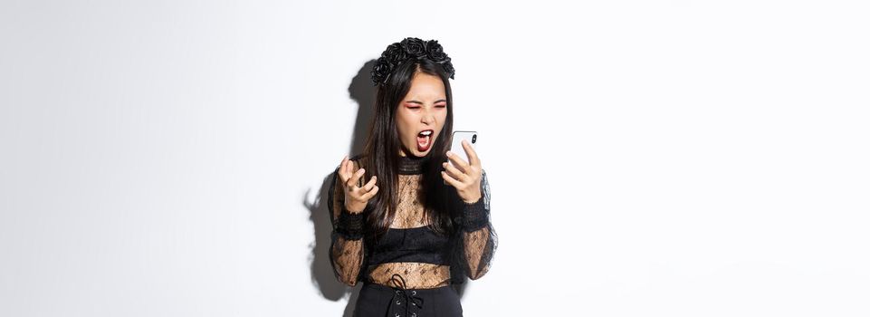 Portrait of angry asian woman in halloween costume looking mad, shouting at mobile phone and grimacing furious, standing over white background.