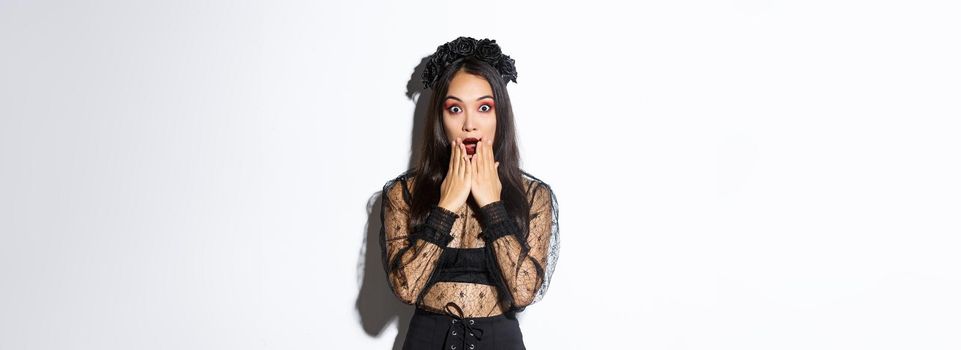 Surprised beautiful asian woman in witch costume looking amazed. Female wearing halloween gothic dress and wreath, open mouth aamused, standing over white background.