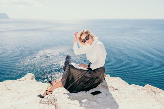 Successful business woman in yellow hat working on laptop by the sea. Pretty lady typing on computer at summer day outdoors. Freelance, travel and holidays concept.