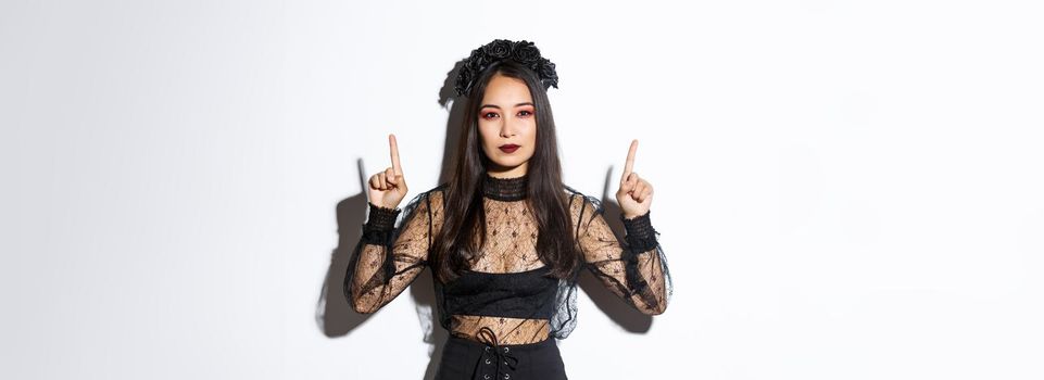 Sassy beautiful asian woman in black gothic dress, wearing witch costume for halloween and pointing fingers up, showing your logo or banner on empty white background, white background.