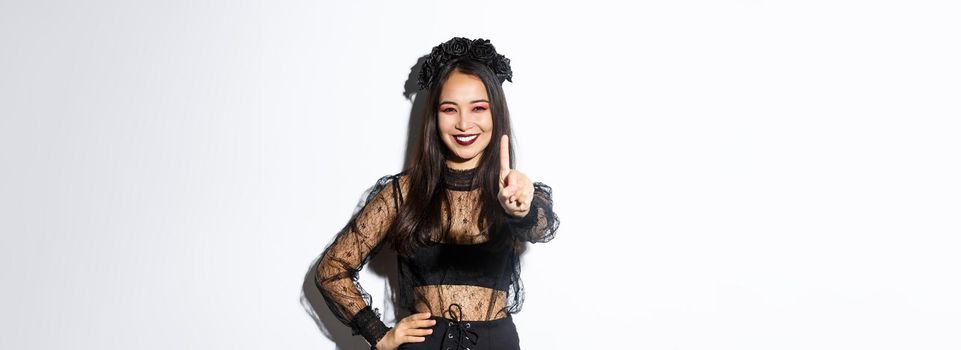 Image of smiling beautiful asian woman in halloween costume showing stop gesture, extend one finger and looking happy, prohibit or reject something, standing white background.