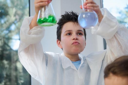 Handsome Caucasian school boy, a scientist chemist in a white lab coat, holding glass laboratory flasks with chemicals, watching the reaction taking place between liquid and reagents. Chemistry class.