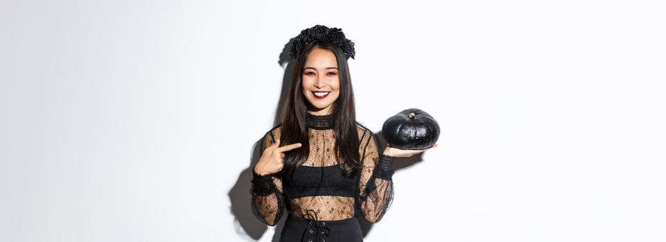 Happy smiling asian woman enjoying halloween celebration, wearing witch costume and pointing finger at black pumpkin, standing over white background.
