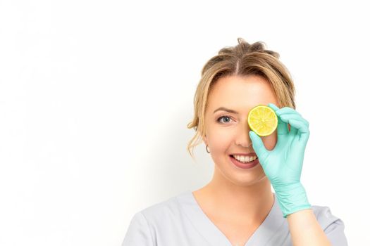 Portrait of young caucasian smiling female beautician covering eye with a lime slice wearing gloves over a white background