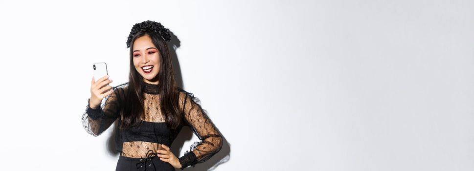 Portrait of happy beautiful asian woman in halloween costume smiling and looking at mobile phone screen, having video call, standing over white background.