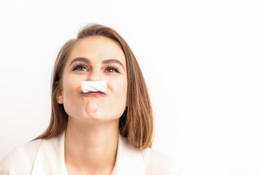 Funny young woman with marshmallow on her lips looking up and standing on white background with copy space