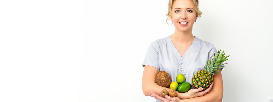 Portrait of a young caucasian smiling female nutritionist with different fruits in her hands over white background