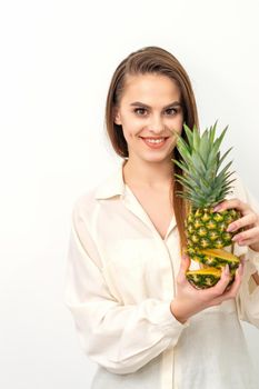 Beautiful young Caucasian woman holding pineapple and smiling, wearing a white shirt over white background