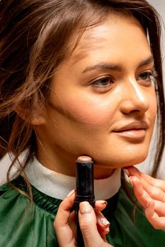 Beautiful young brunette woman receiving makeup with stick concealer on her face in a beauty salon