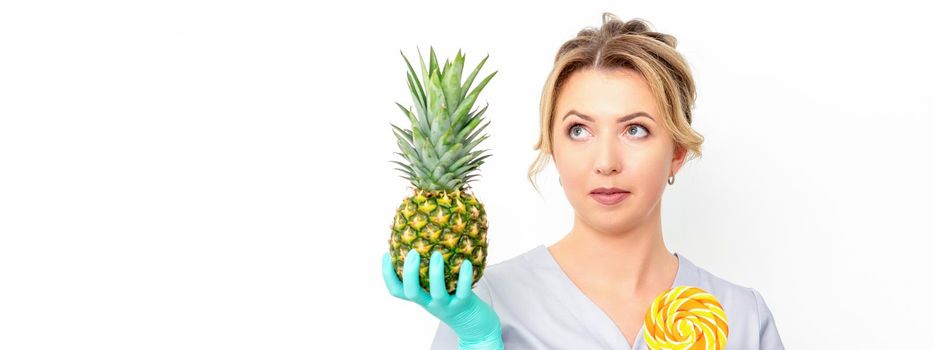 Young caucasian female doctor nutritionist holding fresh pineapple with lollipop over white background