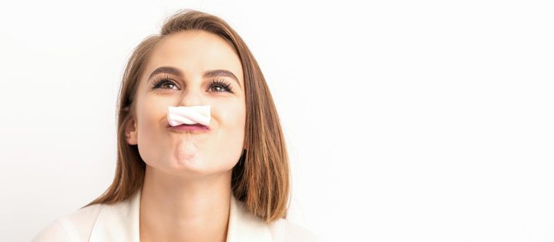 Funny young woman with marshmallow on her lips looking up and standing on white background with copy space