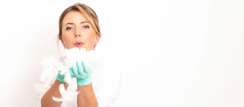 Young beautiful woman beautician in protective green gloves standing and blowing on feathers over white wall background