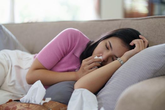 Woman sitting on sofa feeling bad blowing nose sneezing sick.