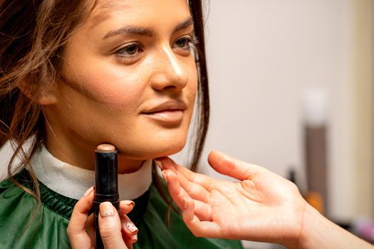 Beautiful young brunette woman receiving makeup with stick concealer on her face in a beauty salon